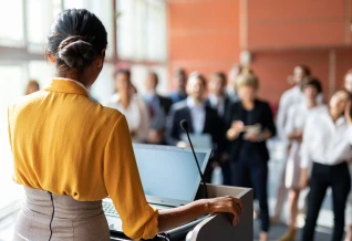
A high-angle photograph showing a woman with her back to the camera, standing at a podium and speaking to a group of people. She is wearing a yellow top and a beige skirt and is holding onto the podium. A laptop is visible on the podium. A crowd of people in professional attire are in the background, facing the speaker. The setting appears to be an indoor business event or a conference.