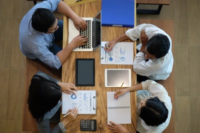 An aerial shot of four people sitting around a wooden conference table, actively engaged in a meeting. They are working with papers, a laptop, a tablet, and a calculator. The image is well-lit and professional, capturing a collaborative and productive work environment.