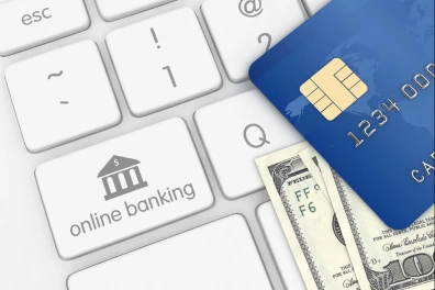  A close-up image of a white computer keyboard. The keys are standard, but one key is customized with an icon of a bank building and the text "online banking". Resting partially on the keyboard is a blue credit card with a gold chip, and underneath the card, the edge of two US dollar bills is visible. The image is clean and clearly represents the concept of online banking, finance, and digital transactions.